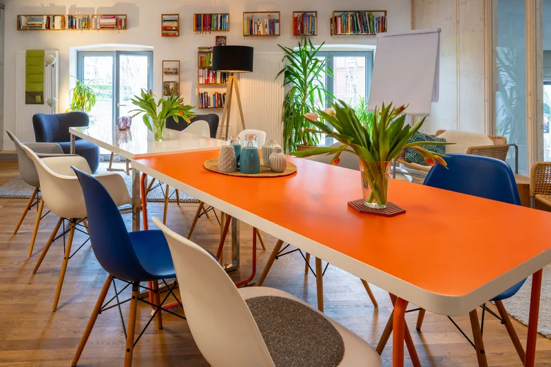 Bücherzimmer der KoFabrik mit orangefarbenem Tisch, Stühlen, Bücherregalen, Pflanzen und Flipchart